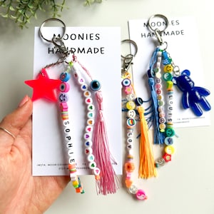 May include: Handmade keychains with beaded accents. One keychain features a neon pink star, the other a blue bear charm. Both have colorful beads, tassels, and the names "SOPHIE" and "LOUIS" spelled out. The keychains are attached to silver keyrings.