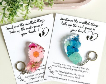 May include: Two resin baby footprint keychains with pressed flowers. The pink keychain has a white daisy and the blue keychain has blue and white flowers. The keychains have the text "Sometimes the smallest things take up the most space in your heart" on the card.