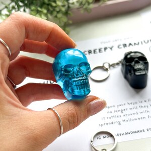 May include: A translucent blue skull keychain is held up, showcasing its intricate details. A black skull keychain is visible in the background. The text "CREEPY CRANIUM" is partially visible, along with other text. The keychains are likely Halloween-themed.
