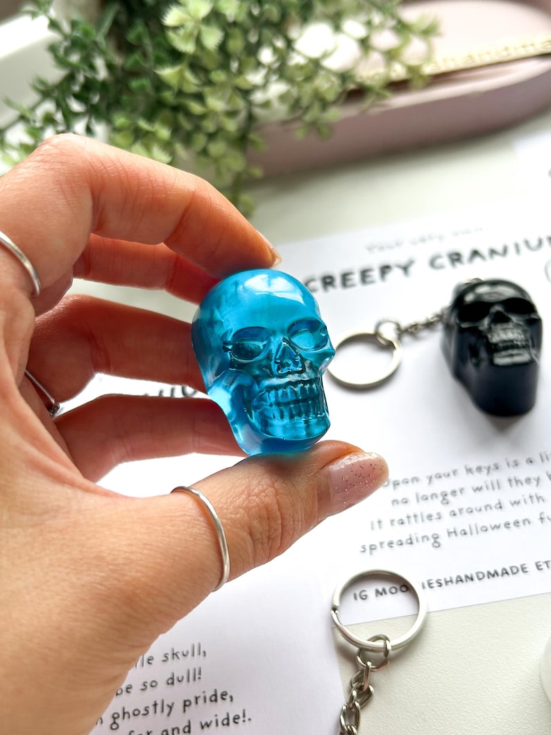 May include: A translucent blue skull keychain is held up, showcasing its intricate details. A black skull keychain is visible in the background. The text "CREEPY CRANIUM" is partially visible, along with other text. The keychains are likely Halloween-themed.