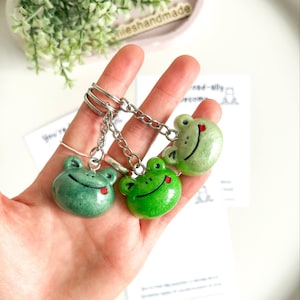 May include: Three green frog keyrings, each with a smiling face and a red tongue. The keyrings are attached to silver chains. The keyrings are held in a hand. The keyrings are in different shades of green.