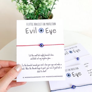 May include: Two cards with blue and white evil eye charms on red and blue string bracelets. The cards have the text "A Little Bracelet for Protection Evil Eye" and "Let this small but mighty bracelet stare, and block out any negative glare..."