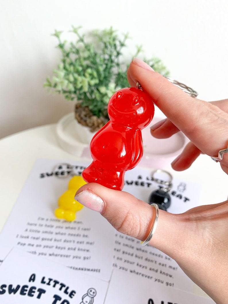 May include: A red gummy bear keychain with a silver key ring. The keychain is being held by a hand. The text "A Little Sweet Treat" is visible in the background.