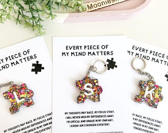 Op de afbeelding: Drie kleurrijke puzzelstukjes sleutelhangers met glitter en de letters L, S en K. Elke sleutelhanger heeft een witte kaart met de tekst "Every piece of my mind matters" en een kort inspirerend bericht.