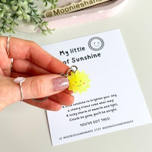 May include: A yellow sun-shaped keychain with a smiling face, attached to a silver chain. The keychain is accompanied by a card with the text "My little of Sunshine" and a poem about the keychain being a lucky charm.