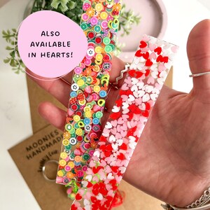 May include: Two clear resin bookmarks with colourful accents. One bookmark features a variety of fruit slices and other shapes. The other bookmark features pink and red hearts. The text "ALSO AVAILABLE IN HEARTS!" is visible in the image.