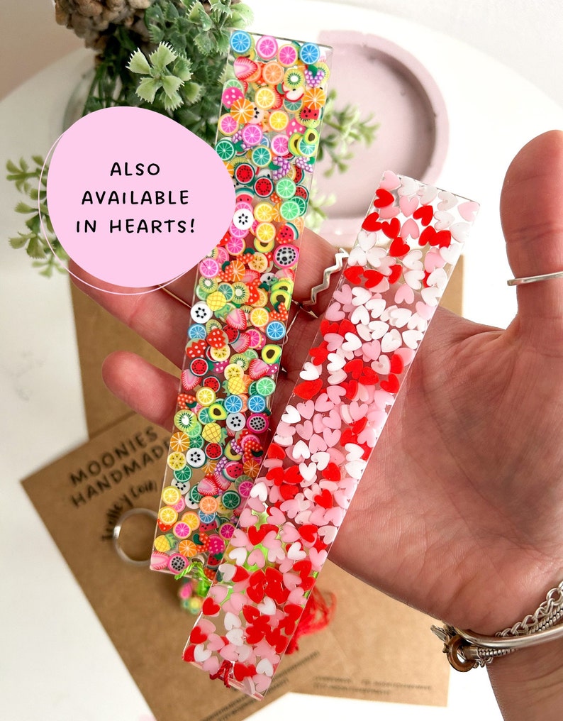 May include: Two clear resin bookmarks with colourful accents. One bookmark features a variety of fruit slices and other shapes. The other bookmark features pink and red hearts. The text "ALSO AVAILABLE IN HEARTS!" is visible in the image.