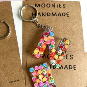 May include: A colourful fruit-themed keychain featuring the letter "A" made from clear resin filled with tiny fruit-shaped charms. The keychain is attached to a silver key ring. The background is a brown paper tag with the text "MOONIES HANDMADE".