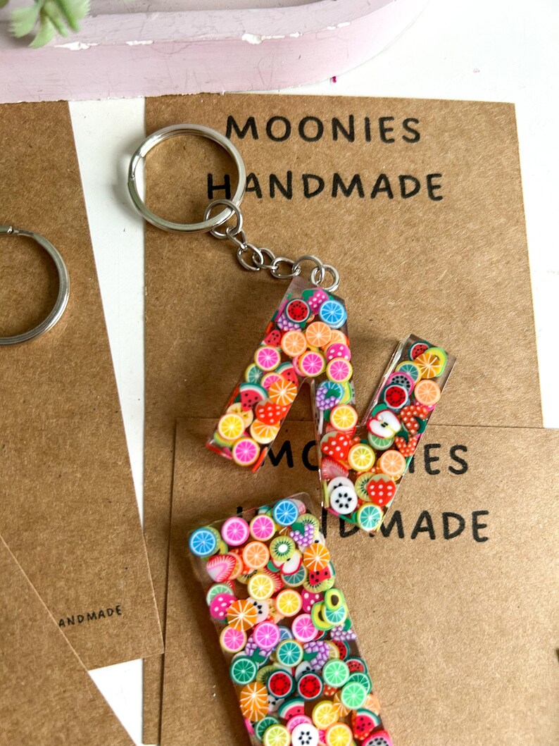 May include: A colourful fruit-themed keychain featuring the letter "A" made from clear resin filled with tiny fruit-shaped charms. The keychain is attached to a silver key ring. The background is a brown paper tag with the text "MOONIES HANDMADE".