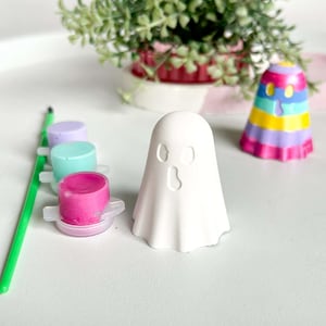 May include: A white ceramic ghost figure with cut-out eyes and mouth, alongside a colourful, striped figure. Paint pots in pink, blue, and purple, and a green paintbrush are also present. A small plant in a pink pot is in the background.