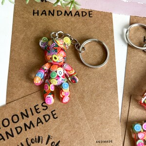 May include: A colourful fruit-themed bear keyring with a silver key ring. The bear is made of polymer clay and has a variety of fruits and sweets on its body. The keyring is handmade and says "Trolley Coin Fab" and "Handmade" on the card.