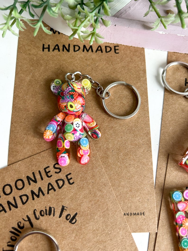 May include: A colourful fruit-themed bear keyring with a silver key ring. The bear is made of polymer clay and has a variety of fruits and sweets on its body. The keyring is handmade and says "Trolley Coin Fab" and "Handmade" on the card.