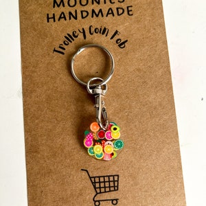 May include: A silver trolley coin fob keyring with a round charm featuring a colourful fruit design. The charm is made of resin and features a variety of fruits, including lemons, limes, oranges, strawberries, and avocados. The keyring is attached to a silver split ring and a silver lobster clasp.