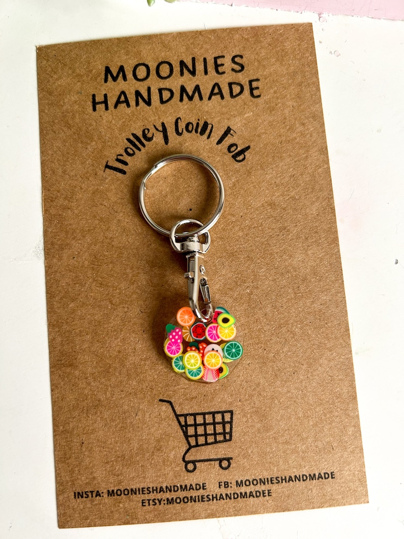 May include: A silver trolley coin fob keyring with a round charm featuring a colourful fruit design. The charm is made of resin and features a variety of fruits, including lemons, limes, oranges, strawberries, and avocados. The keyring is attached to a silver split ring and a silver lobster clasp.