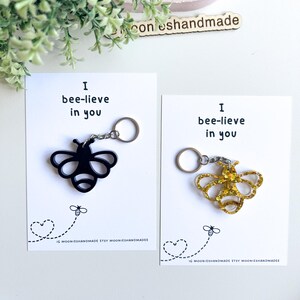 May include: Two bee keyrings, one black and one gold glitter, presented on cards with the text "I bee-lieve in you." Each card features a bee graphic and the Etsy shop name. The keyrings are attached to silver rings.