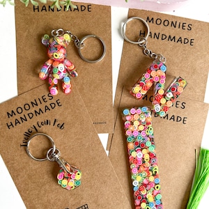 May include: Three handmade fruit-themed keychains. The first keychain is a small bear charm with a colourful fruit design. The second keychain is a letter M charm with a colourful fruit design. The third keychain is a rectangular charm with a colourful fruit design. The charms are attached to silver key rings.