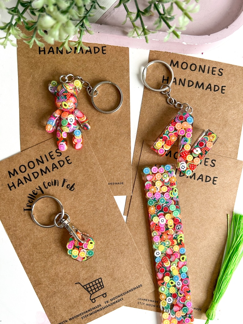 May include: Three handmade fruit-themed keychains. The first keychain is a small bear charm with a colourful fruit design. The second keychain is a letter M charm with a colourful fruit design. The third keychain is a rectangular charm with a colourful fruit design. The charms are attached to silver key rings.