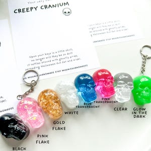 May include: A collection of skull-shaped keychains in various colors, including black, pink, gold flake, white, blue transparent, pink transparent, clear, and glow-in-the-dark green. Each keychain has a metal ring. The text "CREEPY CRANIUM" is visible.