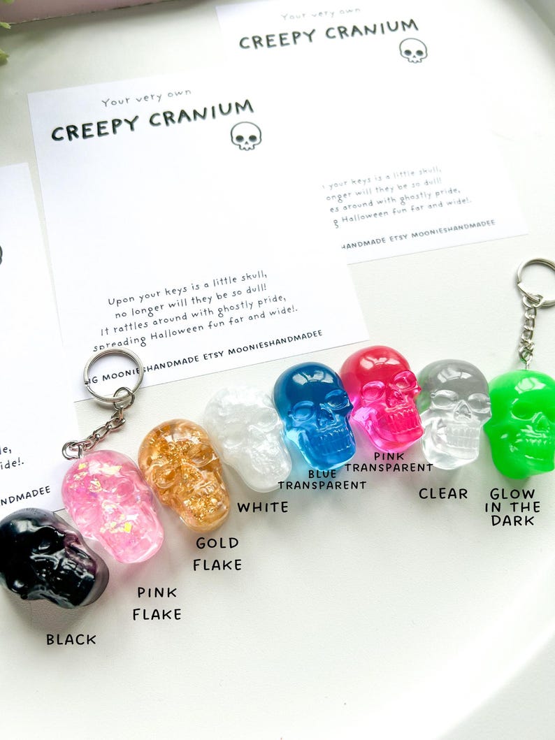 May include: A collection of skull-shaped keychains in various colors, including black, pink, gold flake, white, blue transparent, pink transparent, clear, and glow-in-the-dark green. Each keychain has a metal ring. The text "CREEPY CRANIUM" is visible.