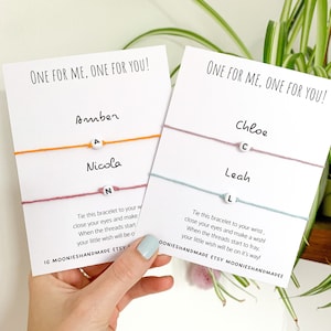 May include: Two white cards with the text "One for me, one for you!" printed on them. Each card has a friendship bracelet with a letter bead attached. The bracelets are orange and pink on one card and blue and light pink on the other card. The letters on the beads are A, N, C, and L.