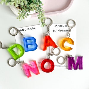May include: Rainbow colored acrylic letter keychains spelling out the word "DOM." The keychains are attached to silver key rings. The keychains are on a white surface with a white card behind them that says "MOONIE HANDMADE."