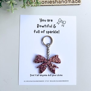 May include: A pink glitter bow keyring with a silver chain, hanging from a white card. The card reads "You are Bowtiful & full of sparkle!" and "Don't let anyone dull your shine."