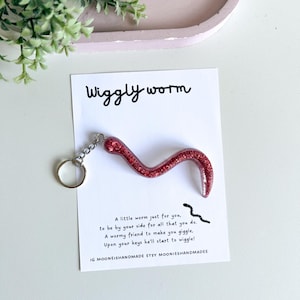 May include: A red glitter worm keyring. The keyring is attached to a white card with the words "Wiggly worm" and a poem. The card is on a white surface. The worm is made of a red glitter material.