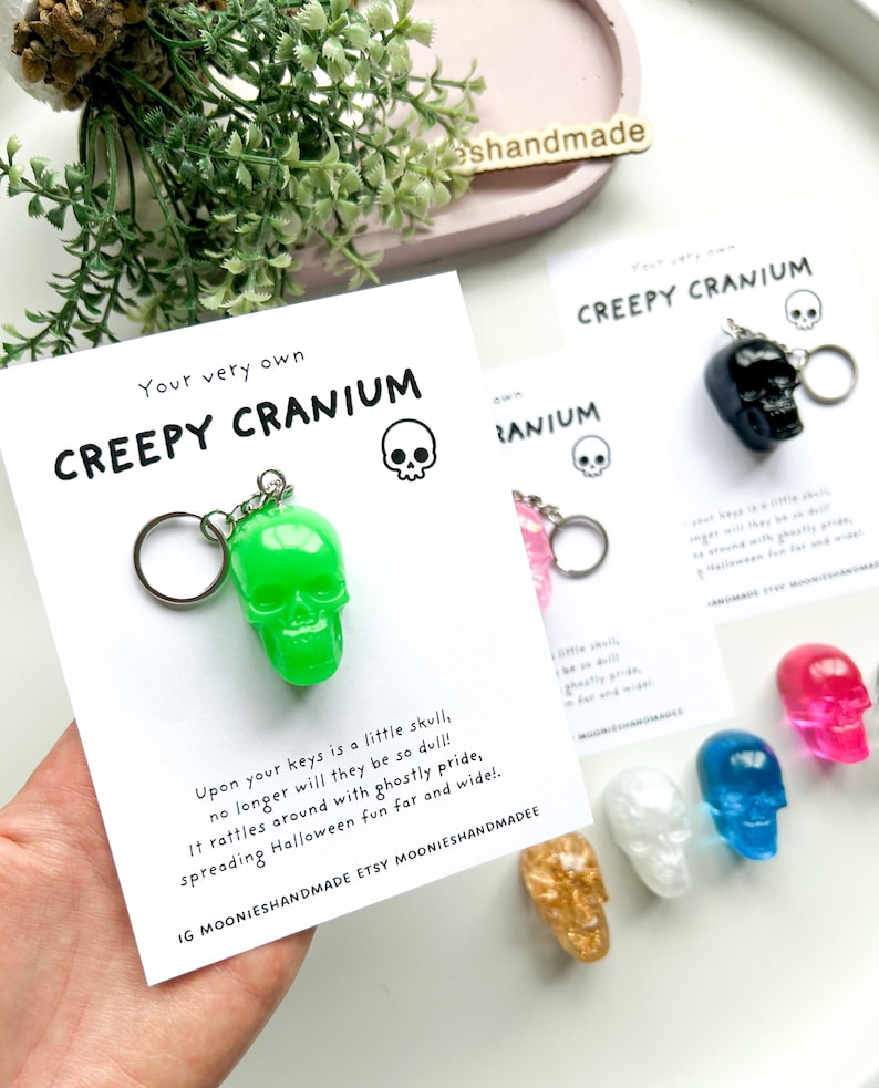May include: A collection of colorful skull keychains. The keychains are in various colors, including green, black, pink, white, blue, gold, and red. Each keychain has a skull design and is attached to a metal ring. The text "CREEPY CRANIUM" is printed on a white card.
