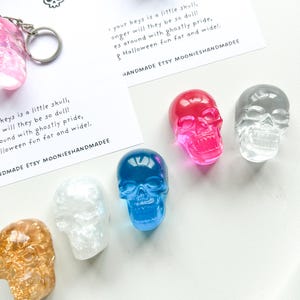 May include: A collection of colorful skull-shaped keychains in various colors, including pink, blue, clear, and gold. The keychains are displayed with a card that reads "your keys is a little skull, longer will they be so dull!" and "Halloween fun far and wide!"