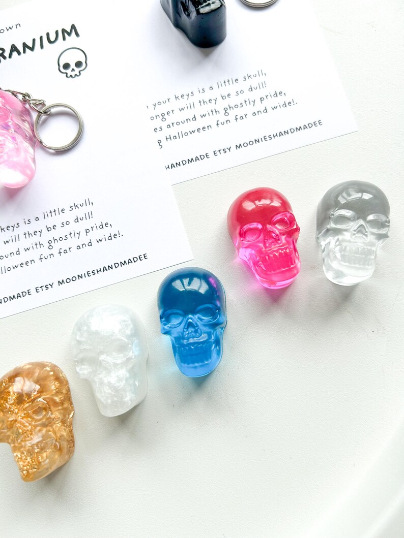 May include: A collection of colorful skull-shaped keychains in various colors, including pink, blue, clear, and gold. The keychains are displayed with a card that reads "your keys is a little skull, longer will they be so dull!" and "Halloween fun far and wide!"