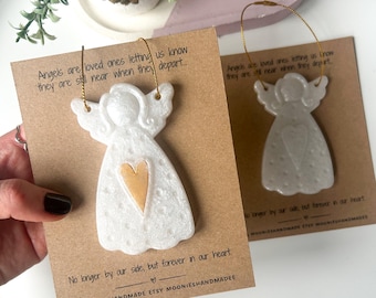 May include: Two white angel ornaments with gold hanging strings, each attached to a brown card. The angels have a gold heart in the center. Text on the cards reads, "Angels are loved ones letting us know they are still near when they depart..." and "No longer by our side, but forever in our heart."