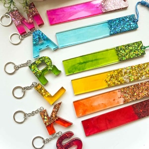 May include: A selection of vibrant resin keyrings and bookmarks. The keyrings are letter-shaped, with silver chain attachments. The bookmarks are rectangular, with glitter detailing. Colours include pink, blue, green, yellow, orange, and red.