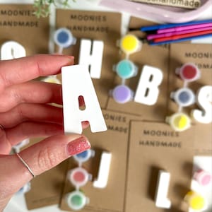 May include: A white letter "A" is held in front of other white letters and paint pots. The letters are part of a DIY paint kit. The background includes the text "Moonies Handmade" on brown cards.