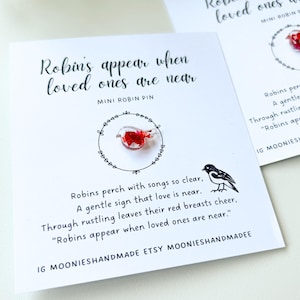May include: Two white cards with the text "Robins appear when loved ones are near" and a mini robin pin. The pin is a small, red bird design. A black and white bird illustration is also present.