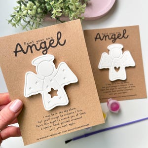 May include: Two craft kits with white angel ornaments. Each kit includes a cardboard backing with the text "Paint your own Angel". One angel has a star cutout, the other a heart. The text reads "I may be in the sky above..."