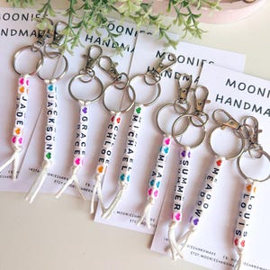 K&ouml;nnte beinhalten: Schl&uuml;sselanh&auml;nger mit Namen aus wei&szlig;en Buchstabenperlen, darunter Jade, Jackson, Grace, Chloe, Michael, Mia, Summer, Meadow und Louis. Die Perlen haben bunte Herzakzente und sind an silbernen Ringen befestigt.