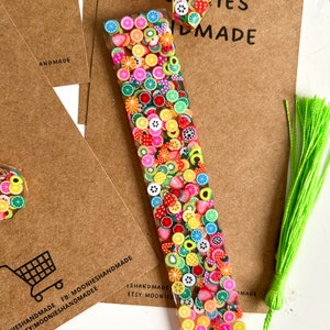 May include: A colourful fruit-themed bookmark with a green tassel. The bookmark is made of clear resin with embedded fruit-shaped charms. The text "MOONIESHANDMADE FB: MOONIESHANDMADE ETSY:MOONIESHANDMADEE" is printed on the brown paper background.
