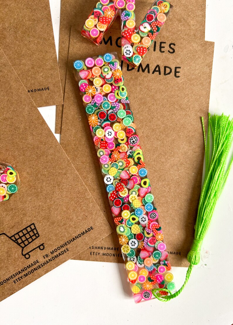 May include: A colourful fruit-themed bookmark with a green tassel. The bookmark is made of clear resin with embedded fruit-shaped charms. The text "MOONIESHANDMADE FB: MOONIESHANDMADE ETSY:MOONIESHANDMADEE" is printed on the brown paper background.