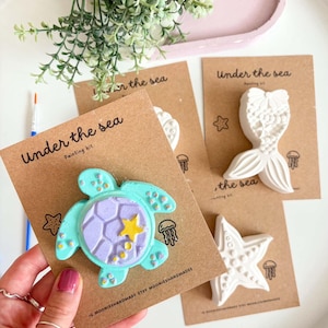 May include: An Under the Sea painting kit featuring a turquoise sea turtle figure with a purple shell and yellow star accent, mounted on a brown card. Additional white plaster figures of a mermaid tail and starfish are displayed on matching cards.
