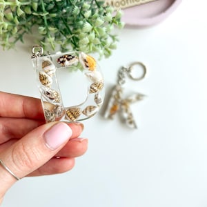 May include: Clear resin letter "D" and "K" keychains filled with seashells. The "D" is held by a hand. The keychains have silver-colored hardware. The background includes greenery and a pink tray with the text "Moonieshandmade".