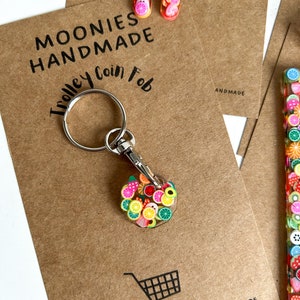 May include: A colourful fruit-themed keychain with a silver keyring and a clip. The keychain is made of polymer clay and features a variety of fruits, including strawberries, lemons, oranges, and limes. The keychain is labelled "Trolley Coin Fob" and "Moonies Handmade".