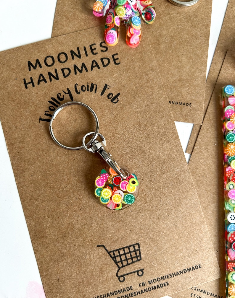 May include: A colourful fruit-themed keychain with a silver keyring and a clip. The keychain is made of polymer clay and features a variety of fruits, including strawberries, lemons, oranges, and limes. The keychain is labelled "Trolley Coin Fob" and "Moonies Handmade".