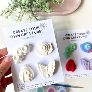May include: A pottery painting set with the text "CREATE YOUR OWN CREATURES". The set includes unpainted ceramic creatures, paint pots, and a paintbrush. The creatures include a snail, butterfly, and ladybug. Ideal for creative activities.