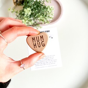 May include: A wooden heart-shaped token with the words "MUM HUG" engraved on it. The token is being held by a hand.