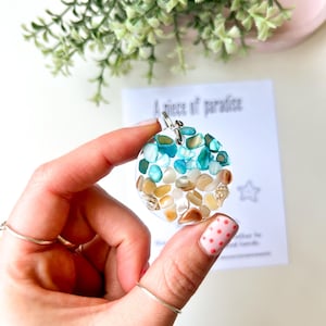 May include: A circular pendant with a clear resin base, filled with small, colourful stones in shades of blue, green, tan, and white. The pendant has a silver-coloured bail. The text "A piece of paradise" is visible on a card in the background.