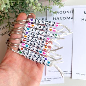 K&ouml;nnte beinhalten: Eine Sammlung personalisierter Schl&uuml;sselanh&auml;nger mit Namen, die in wei&szlig;en Buchstabenperlen geschrieben sind. Jeder Schl&uuml;sselanh&auml;nger hat einen anderen Namen, akzentuiert mit bunten Herzperlen. Die Schl&uuml;sselanh&auml;nger sind an silbernen Schl&uuml;sselringen und wei&szlig;en Kordeln befestigt.