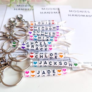 K&ouml;nnte beinhalten: Schl&uuml;sselanh&auml;nger mit Namen, die in wei&szlig;en Buchstabenperlen geschrieben sind, akzentuiert mit bunten Herzperlen. Namen sind Louis, Meadow, Summer, Mia, Michael, Grace, Chloe, Jackson und Jade. Jeder Schl&uuml;sselanh&auml;nger hat einen silbernen Verschluss.