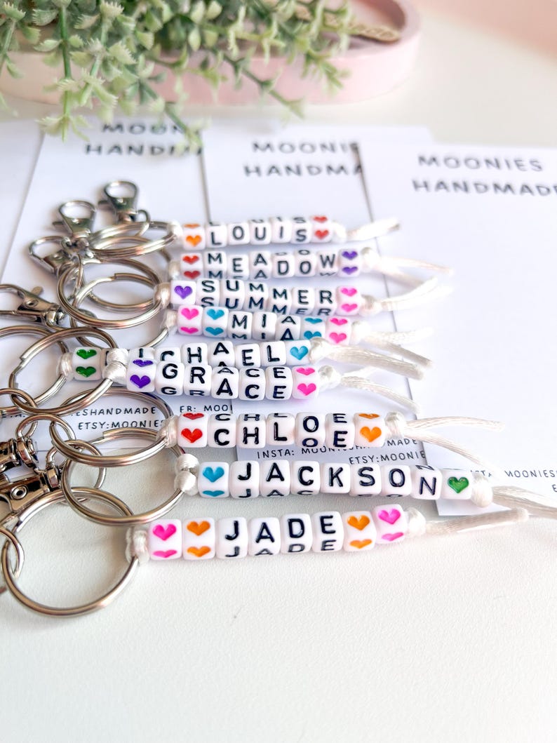 K&ouml;nnte beinhalten: Schl&uuml;sselanh&auml;nger mit Namen, die in wei&szlig;en Buchstabenperlen geschrieben sind, akzentuiert mit bunten Herzperlen. Namen sind Louis, Meadow, Summer, Mia, Michael, Grace, Chloe, Jackson und Jade. Jeder Schl&uuml;sselanh&auml;nger hat einen silbernen Verschluss.