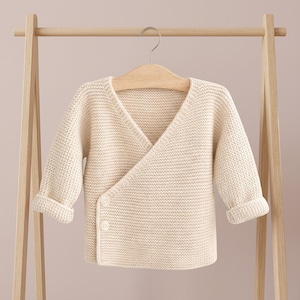 Könnte beinhalten: Cremefarbener Baby-Wickelpullover mit langen Ärmeln und V-Ausschnitt. Der Pullover hat ein Wickeldesign mit zwei Knöpfen zum Verschließen. Er hängt an einem Holzkleiderbügel auf einem hölzernen Kleiderständer.
