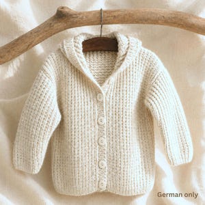 Pode incluir: Cardigã de bebê com capuz na cor creme, com mangas compridas e fecho de botões. O suéter de malha é exibido em um cabide de madeira, destacando sua textura macia e design. A cor neutra o torna adequado para várias roupas.